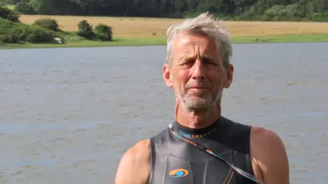 BBC Ric Cooper of The Cleddau Project ready to go for a swim in the Western Cleddau river. He's standing in front of the river wearing a black, sleeveless wetsuit. He has grey short hair and grey stubble.