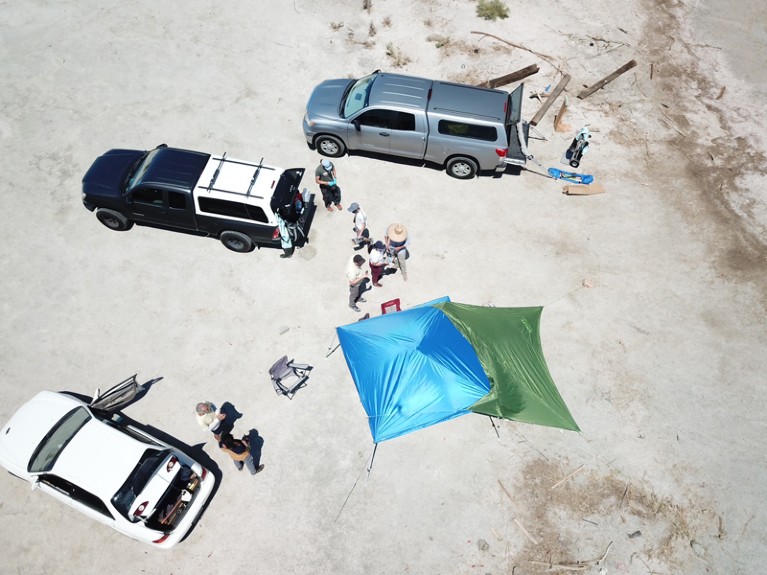 Aerial overview of cars and tents pitched on a dry lake shore and field researchers working in bright sunshine