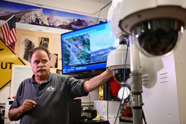 A man gestures towards a set of two surveillance cameras and a screen behind him shows satellite photos with data points overlaid on them
