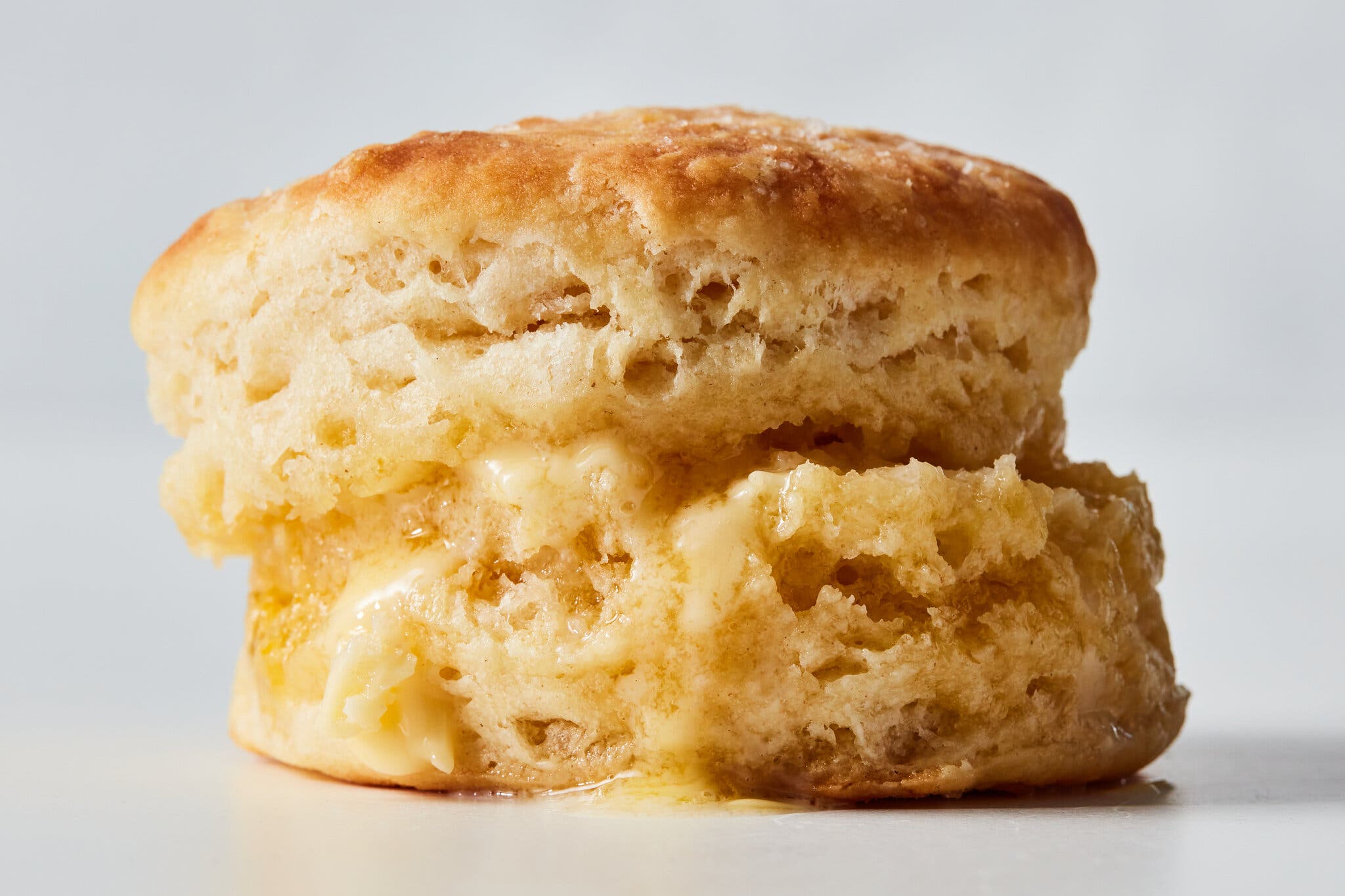 A fluffy, golden biscuit stands on a light surface, with melting butter dripping from its middle.
