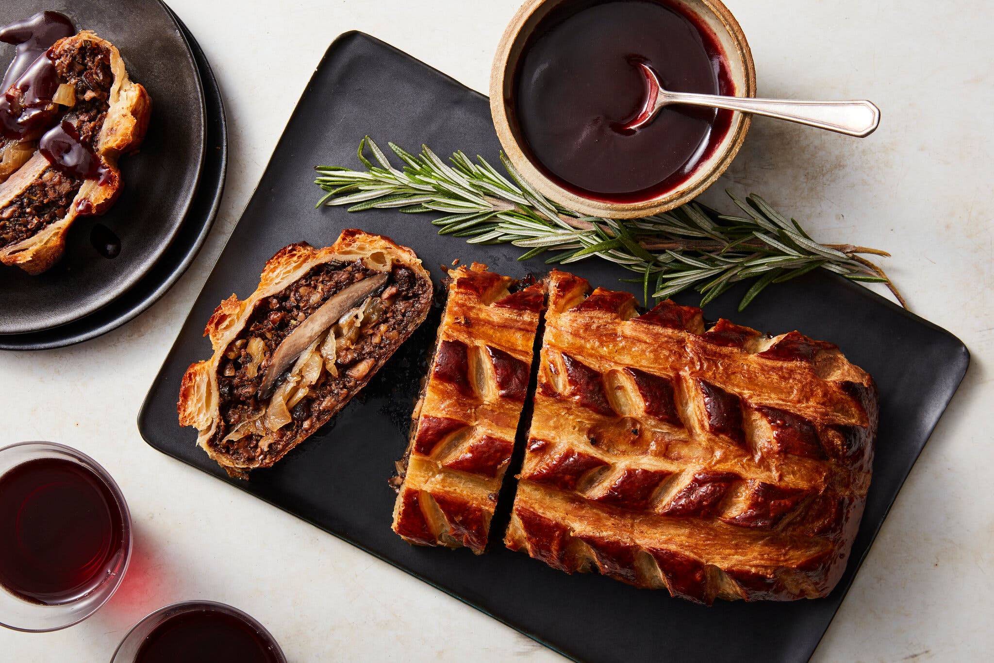 A sliced, golden-brown Wellington pastry with a mushroom and onion filling rests on a black platter. Dark red sauce, rosemary, and wine glasses sit nearby.