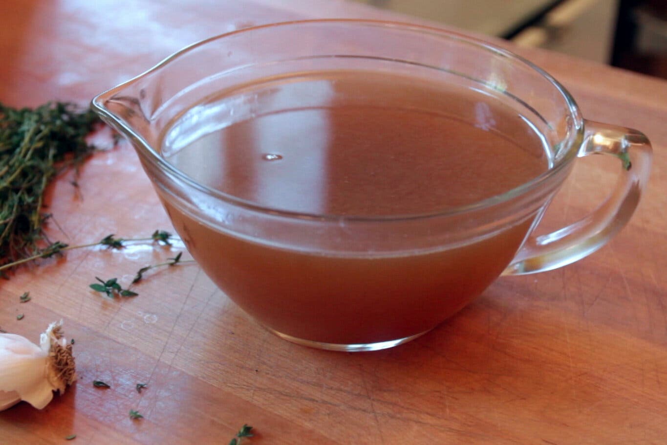 A glass bowl of turkey stock sits on a wooden counter, with herbs and a head of garlic nearby.