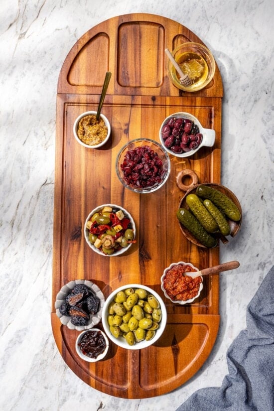 Bowls of spreads, pickles, and olives on wooden charcuterie board.