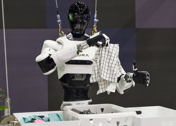 Humanoid robot with black visor folds laundry, holding checkered towel beside basket, suspended in lab with cables and equipment.