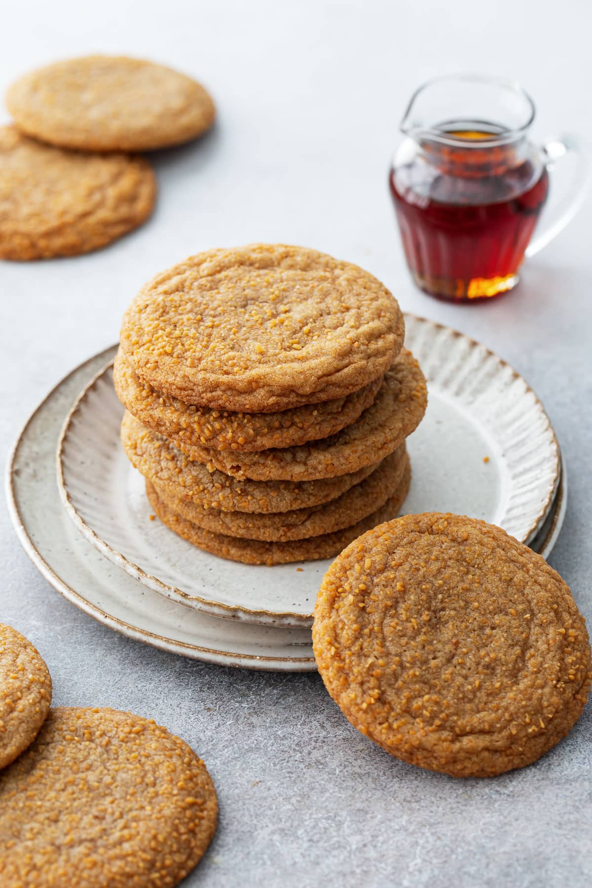 Stack of Chewy Maple Brown Butter Sugar Cookies on a ceramic plate, with a small glass pitcher of maple syrup and more cookies scattered around.