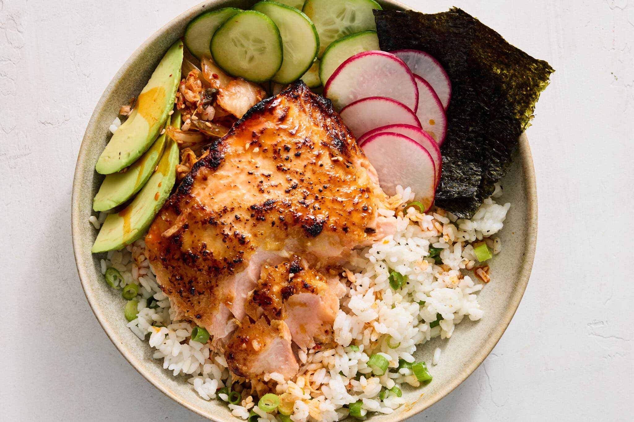 An overhead image of a rice bowl topped with salmon, sliced avocado, cucumbers and radishes.